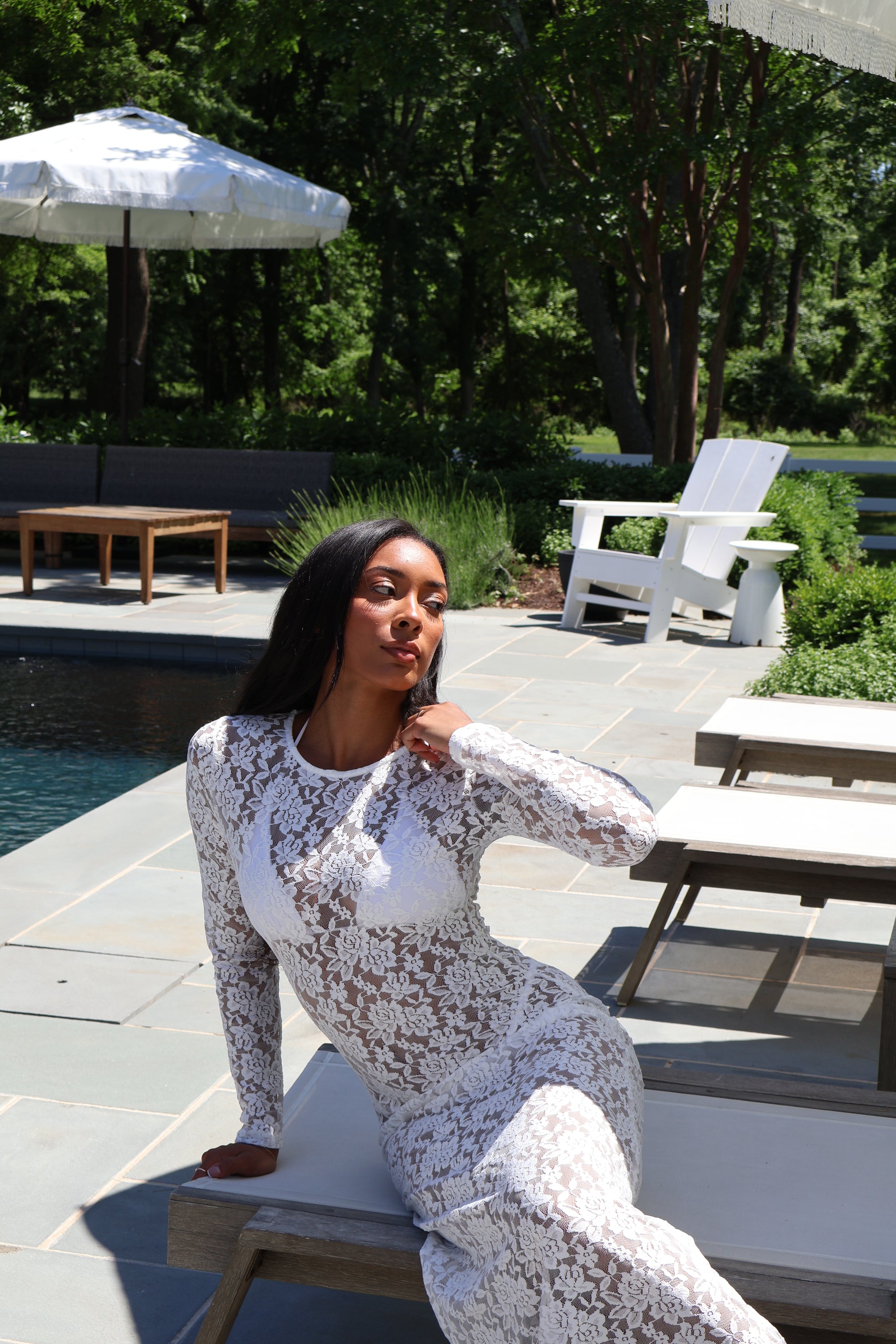 Woman in a white lace dress sitting by a poolside with greenery in the background